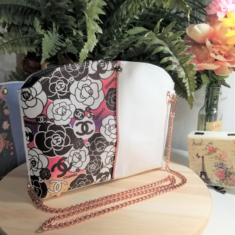 May include: A white and black floral print handbag with a rose pattern and the word "Chanel" repeated throughout. The bag has a white leather flap and a rose gold chain strap.