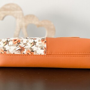 May include: A small orange and white floral patterned clutch purse with a zipper closure.