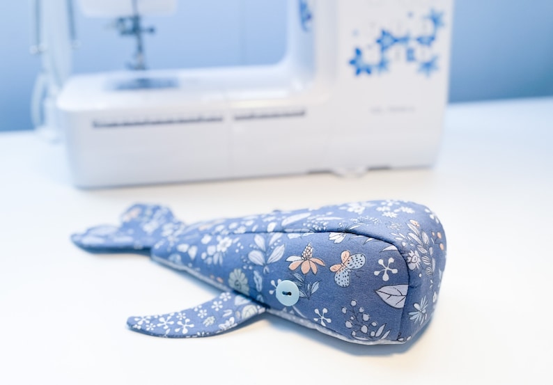 Puede incluir: Un juguete de peluche con forma de ballena azul con un patr&oacute;n floral y un bot&oacute;n azul claro. El juguete est&aacute; acostado sobre una superficie blanca.