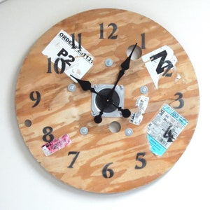 May include: A wooden clock with black numbers and hands. The clock face is made from a wooden spool with various labels and stickers on it. The clock is hanging on a white wall.