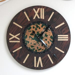 May include: A round wooden clock with Roman numerals. The clock face is dark brown with light beige Roman numerals and hands. The center of the clock has a green and tan pattern, surrounded by black stones.