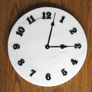 May include: A white marble clock face with black numbers and hands. The clock face is round and has a diameter of approximately 12 inches.