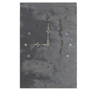 May include: A slate clock face with a gold-colored hour and minute hand. The clock face has a rustic, weathered look with a crack running through it. There are 12 small holes around the edge of the clock face, where the numbers would be.