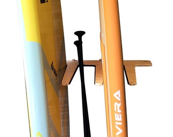 Puede incluir: Un soporte de pared de madera para tablas de paddle surf con dos tablas de paddle surf y dos palas. Las tablas de paddle surf son naranja y azul con detalles amarillos. Las tablas de paddle surf tienen las palabras "Riviera" y "Rider" impresas en ellas.
