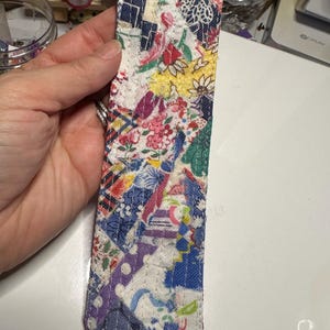 May include: A colourful fabric bookmark with a patchwork design. The bookmark is made of different fabrics with floral and geometric patterns in shades of blue, red, yellow, green, and purple.