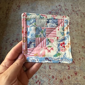 Puede incluir: Un pequeño posavasos cuadrado de patchwork con una variedad de cuadrados de tela estampados con flores y rayas en tonos azules, rojos, rosas y blancos. Los bordes están acabados con una tela floral rosa y un borde floral azul. El posavasos se sostiene en una mano.