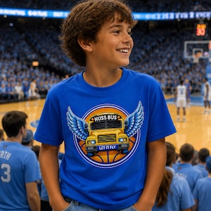 Könnte beinhalten: Ein blaues T-Shirt mit einem gelben Schulbus-Design, blauen Flügeln und dem Text "HUSS BUS LET IT FLY". Das T-Shirt wird von einer Person in einer Basketballarena getragen.