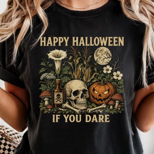 May include: A black t-shirt featuring a graphic print with the text HAPPY HALLOWEEN IF YOU DARE. The design includes a human skull, a jack-o'-lantern, a bottle with a skull and crossbones label, mushrooms, corn stalks, and a full moon.