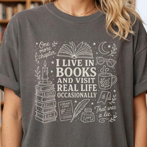 Blonde woman wearing a Pepper garment-dyed T-shirt in a bookstore. The cream doodle design reads “I Live in Books and Visit Real Life Occasionally” with books, an open book, glasses, a mug, moon, stars, candle, and ink bottle.