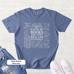 Blue Jean garment-dyed T-shirt shown flat with pale book-themed doodle art reading “I Live in Books and Visit Real Life Occasionally,” with books, glasses, coffee mug, candle, moon, stars, and literary illustrations.