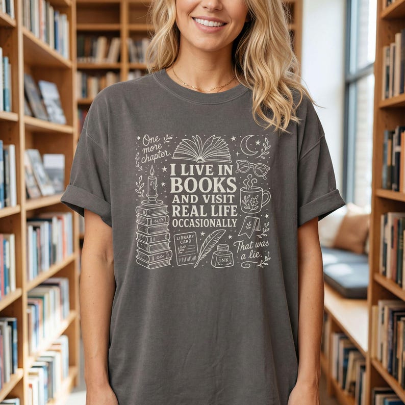 Blonde woman wearing a Pepper garment-dyed T-shirt in a bookstore. The cream doodle design reads “I Live in Books and Visit Real Life Occasionally” with books, an open book, glasses, a mug, moon, stars, candle, and ink bottle.