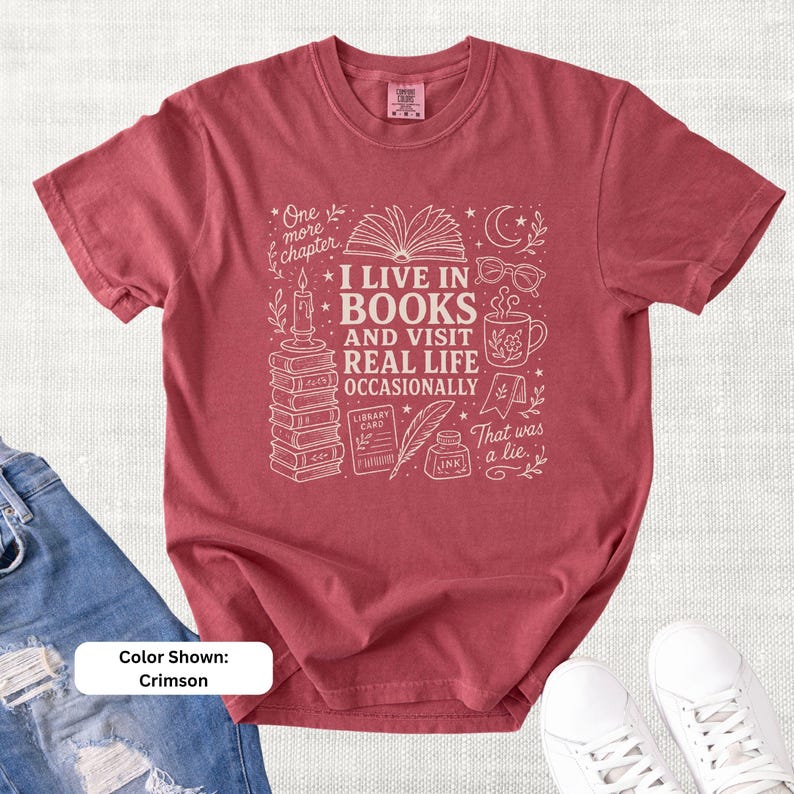 Crimson red garment-dyed T-shirt shown flat with cream book-themed doodle art reading “I Live in Books and Visit Real Life Occasionally,” surrounded by stacked books, an open book, glasses, a mug, candle, moon, stars, and ink bottle.