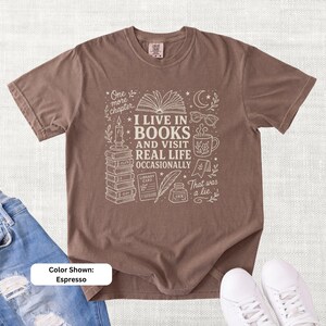 Espresso brown garment-dyed T-shirt shown flat with cream book-lover doodle art reading “I Live in Books and Visit Real Life Occasionally,” surrounded by books, glasses, a mug, moon, stars, and reading icons.