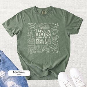 Moss green garment-dyed T-shirt shown flat with a cream doodle design reading “I Live in Books and Visit Real Life Occasionally,” featuring stacked books, an open book, glasses, a mug, candle, moon, stars, and ink bottle.