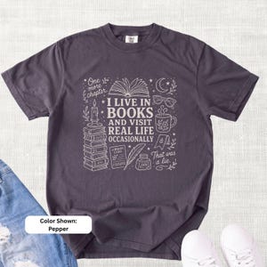 Pepper garment-dyed T-shirt shown flat on a light linen background with jeans and white sneakers. Cream book-themed doodle art reads “I Live in Books and Visit Real Life Occasionally.”