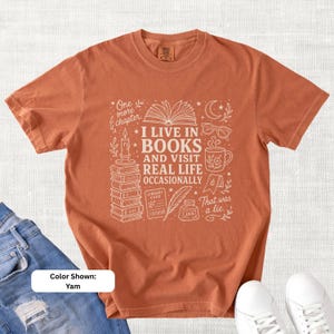 Yam orange garment-dyed T-shirt shown flat with pale bookish doodle artwork reading “I Live in Books and Visit Real Life Occasionally,” featuring books, glasses, a coffee mug, candle, moon, stars, and reading illustrations.