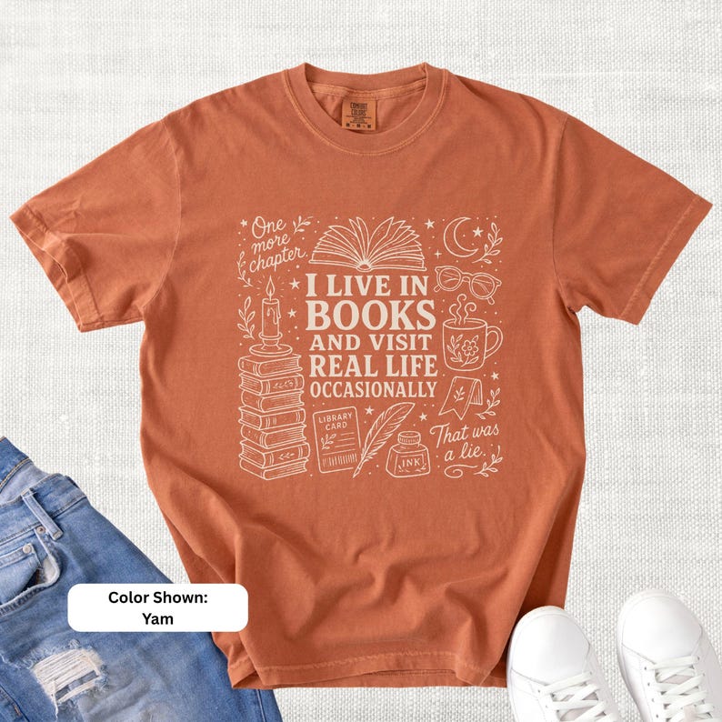 Yam orange garment-dyed T-shirt shown flat with pale bookish doodle artwork reading “I Live in Books and Visit Real Life Occasionally,” featuring books, glasses, a coffee mug, candle, moon, stars, and reading illustrations.