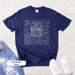 Navy garment-dyed T-shirt shown flat with cream bookish doodle artwork reading “I Live in Books and Visit Real Life Occasionally,” surrounded by stacked books, an open book, glasses, a mug, candle, moon, stars, and ink bottle.
