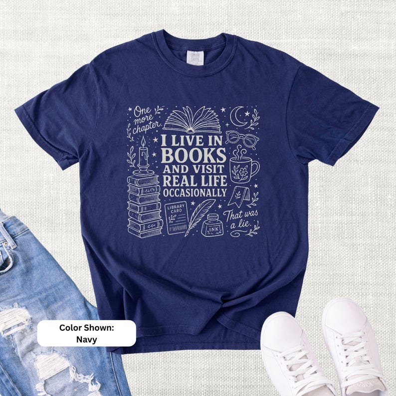 Navy garment-dyed T-shirt shown flat with cream bookish doodle artwork reading “I Live in Books and Visit Real Life Occasionally,” surrounded by stacked books, an open book, glasses, a mug, candle, moon, stars, and ink bottle.