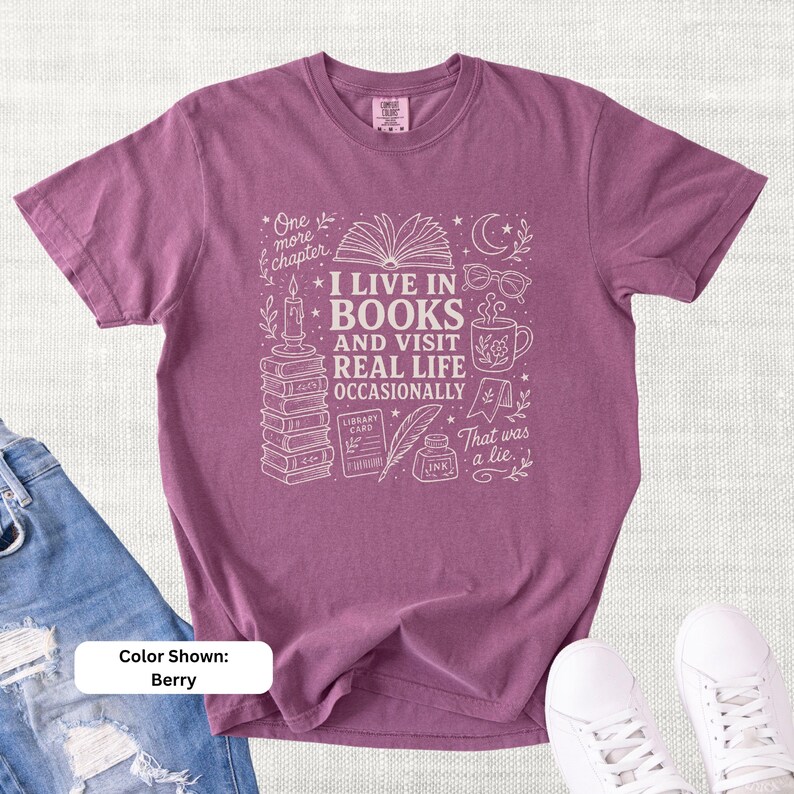 Berry purple garment-dyed T-shirt shown flat with pale book-lover doodle art reading “I Live in Books and Visit Real Life Occasionally,” featuring books, glasses, a coffee mug, candle, moon, stars, and literary icons.