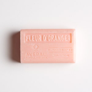 May include: A rectangular, light pink bar of soap with the words "FLEUR D'ORANGER" embossed on the top. The soap also features the brand name "AVERAL" and the text "MADE IN FRANCE".