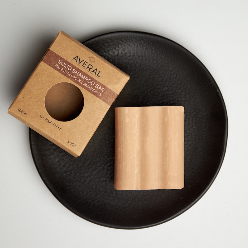 Op de afbeelding: Een lichtbruine vaste shampoo bar met een geribbeld oppervlak ligt op een zwart bord naast de kartonnen verpakking. De verpakking leest "AVERAL SOLID SHAMPOO BAR MADE WITH ORGANIC INGREDIENTS" en "ALL HAIR TYPES". De bar weegt 100g.