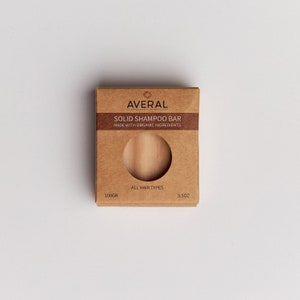 Op de afbeelding: Een bruine kartonnen doos met een vaste shampoo bar. De doos heeft de merknaam "AVERAL" en de tekst "SOLID SHAMPOO BAR MADE WITH ORGANIC INGREDIENTS". De shampoo bar is zichtbaar door een cirkelvormige uitsparing. Het weegt 100g en is voor alle haartypes.