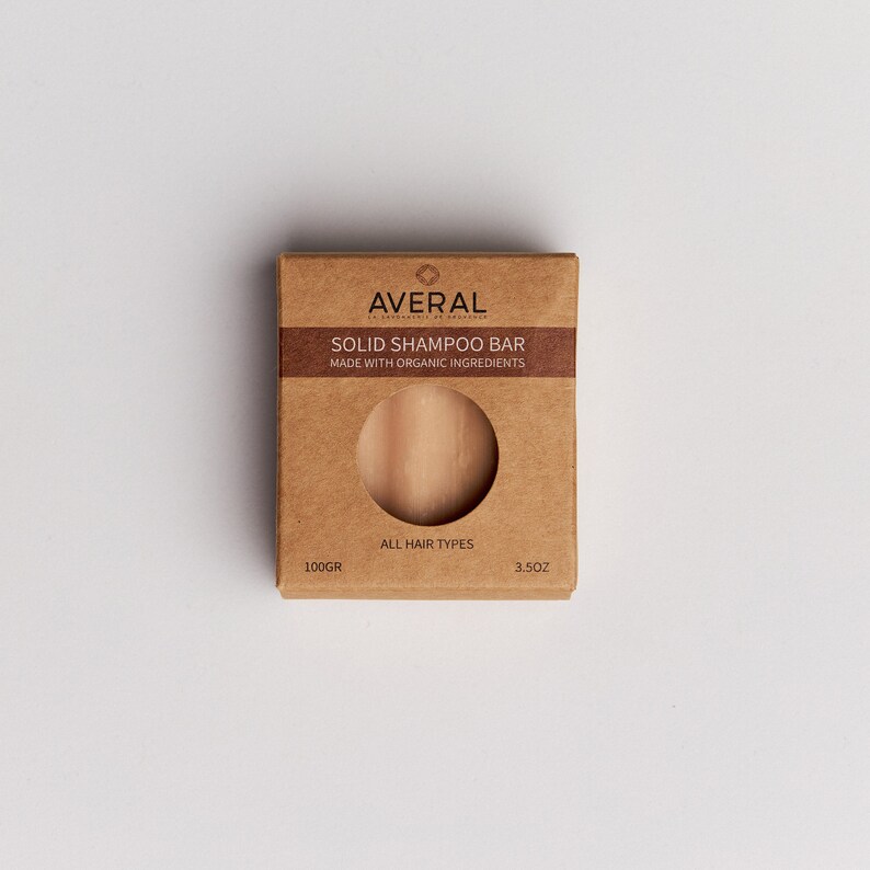 Op de afbeelding: Een bruine kartonnen doos met een vaste shampoo bar. De doos heeft de merknaam "AVERAL" en de tekst "SOLID SHAMPOO BAR MADE WITH ORGANIC INGREDIENTS". De shampoo bar is zichtbaar door een cirkelvormige uitsparing. Het weegt 100g en is voor alle haartypes.