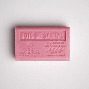 May include: A pink bar of soap with the text "BOIS DE SANTAL" in white lettering. The soap also has the text "AU BEURRE DE KARITE BIO" and "AVERAL SAVON DE PROVENCE MADE IN FRANCE" in white lettering.