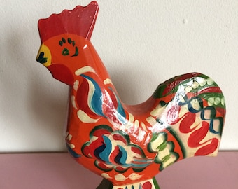 May include: A hand-painted wooden rooster figurine with a red, orange, green, blue, and white floral design. The rooster is standing on a base with a similar floral design.