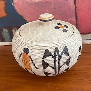 May include: A white woven basket with a lid, featuring black and orange geometric designs. The basket has a round shape and a textured surface.
