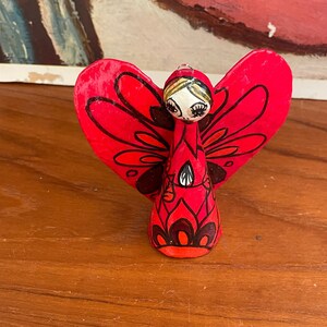 May include: A red and black angel ornament with a heart-shaped body and wings. The angel has a black and white face with large eyes and a black hair bow.
