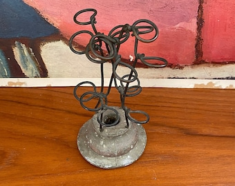 May include: A vintage metal candle holder with a circular base and an abstract wire design. The candle holder has a weathered, aged appearance, with a dark, metallic finish. The design features looping wire elements that create an artistic, sculptural form.