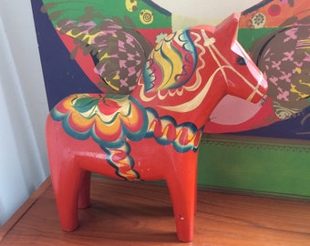 Pode incluir: Um cavalo Dala sueco tradicional, um brinquedo de madeira pintado à mão, é vermelho com desenhos florais coloridos. O cavalo está em pé sobre uma superfície de madeira.