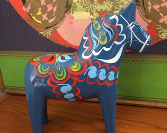Puede incluir: Un caballo de madera azul con diseños pintados de colores. El caballo tiene una crin y una cola blancas y está decorado con pintura roja, amarilla, verde y azul.