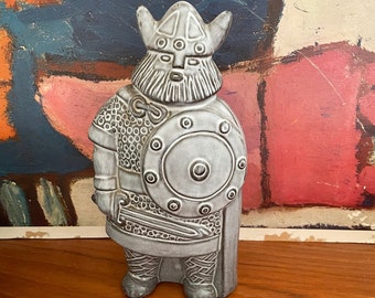 May include: A gray ceramic figurine of a Viking warrior wearing a helmet with horns and holding a shield and sword.