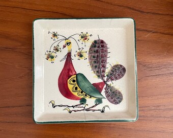 May include: A white ceramic square dish with a green border. The dish features a hand-painted design of a stylized bird with yellow, red, and green colors.
