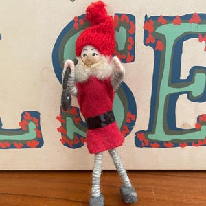 May include: A handmade Christmas elf figurine. The elf wears a red felt outfit with a black belt, a red knit hat, and gray shoes. The elf has a white beard and is holding a black purse. The background is a light beige color with teal and red accents.