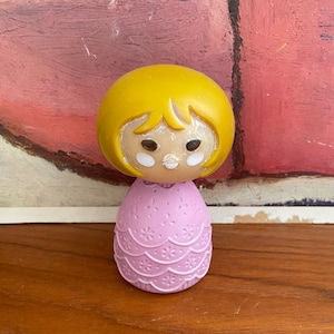 May include: A small plastic doll with a yellow head, brown eyes, and a pink dress with a white floral pattern.