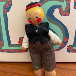 May include: A vintage wooden doll with a painted yellow head, rosy cheeks, and blue eyes. The doll wears a black vest, brown pants, and a small hat with a red band. The doll's arms are made of a light-colored fabric.