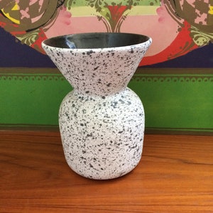 May include: A white ceramic vase with black speckles and a black rim. The vase has a wide mouth and a narrow base.