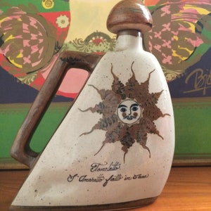 May include: A ceramic bottle with a brown and white glaze. The bottle has a handle and a cork stopper. The bottle is decorated with a sun with a face and the text "Toranietto S. Amaretto fatto in casa".