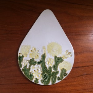 May include: White ceramic trivet in the shape of a teardrop with a raised design of yellow flowers and green leaves.
