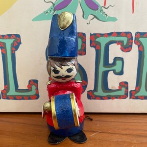 May include: A miniature wooden toy soldier figurine. The figurine is painted with a blue hat, red jacket, and black boots. It holds a blue and gold drum. The soldier has a painted face with a mustache.