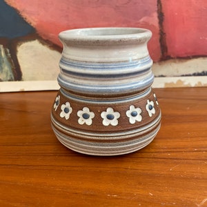 May include: A small, brown and white ceramic vase with a blue and white striped pattern and a row of white flowers with blue centers around the middle.
