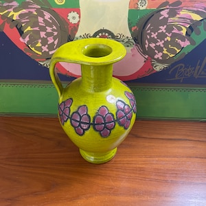 May include: A green ceramic vase with a handle and a floral design. The vase has a textured surface and a wide mouth.