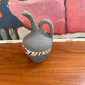 May include: A small gray ceramic pitcher with a handle and a decorative band of red, yellow, blue, and white waves around the middle.
