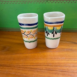 May include: Two white ceramic shot glasses with colorful hand-painted designs. One glass features a green, yellow, and orange floral pattern. The other glass has a green, yellow, and black geometric pattern.