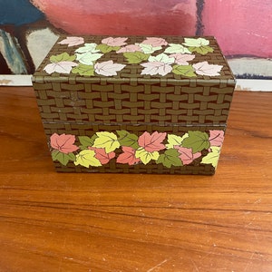 May include: A rectangular brown metal box with a woven basket pattern. The box is decorated with colorful leaves in shades of pink, yellow, and green. The box is sitting on a wooden surface.
