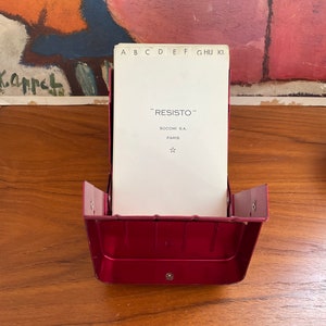 May include: A red metal card holder with a white card inside. The card has the text "RESISTO" SOCOMI S.A. PARIS and a star.